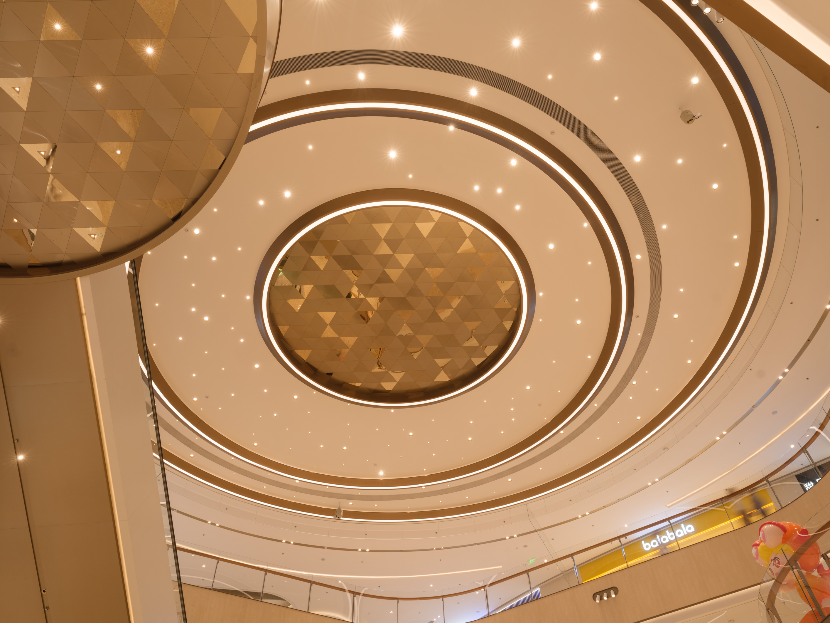 购物中心内现代圆形天花板，中央区域有三角形镜面装饰，周围环绕暖色灯光。